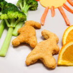 Dino nuggets on a gray plate next to broccoli trees, a cheese and carrot sun, and orange slices.