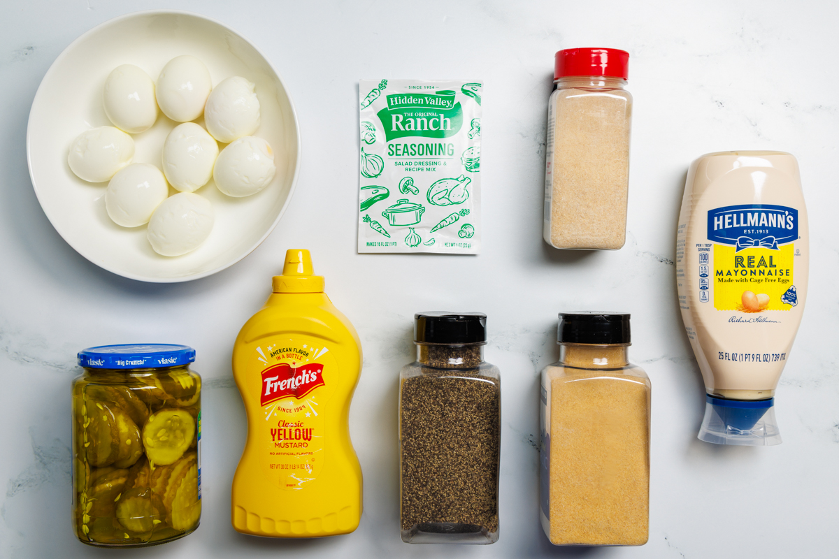 Dill Pickle Egg Salad recipe ingredients in various containers on a white marble surface: hard boiled eggs (peeled and dried), ranch seasoning mix, onion powder, mayonnaise, dill pickles, dill pickle juice, yellow mustard, ground black pepper, and garlic powder.