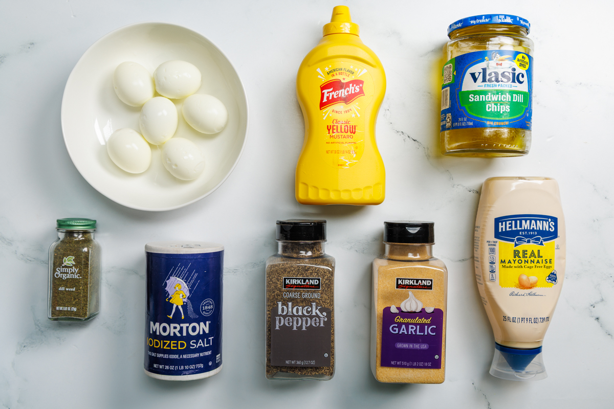 Dill Pickle Deviled Eggs recipe ingredients in various containers on a white marble surface: hard boiled eggs (cooled and peeled), yellow mustard, dill pickle chips, dill pickle juice, dill weed, salt, ground black pepper, garlic powder, and mayonnaise.