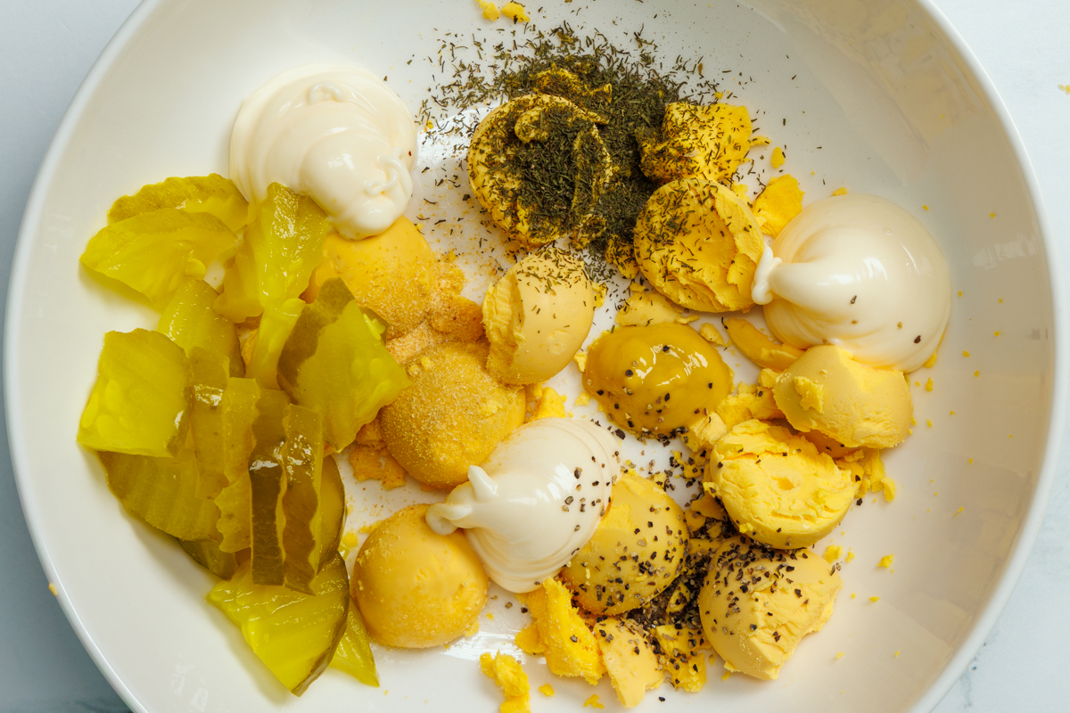 Hard boiled egg yolks, mayonnaise, yellow mustard, sliced dill pickle chips, dill pickle juice, dried dill weed, salt, ground black pepper, and garlic powder in a white bowl.