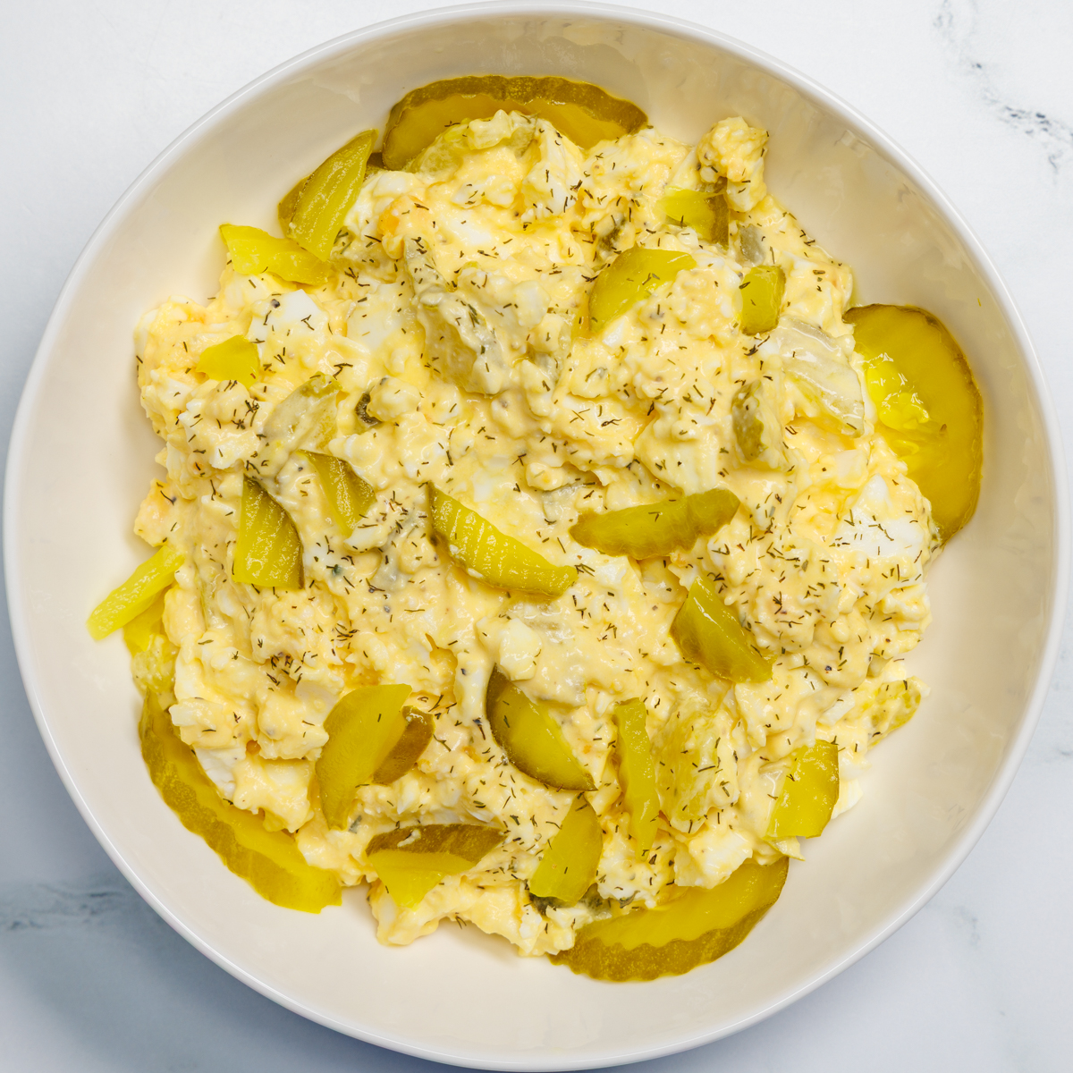 Dill pickle egg salad in a white bowl on a white marble surface.