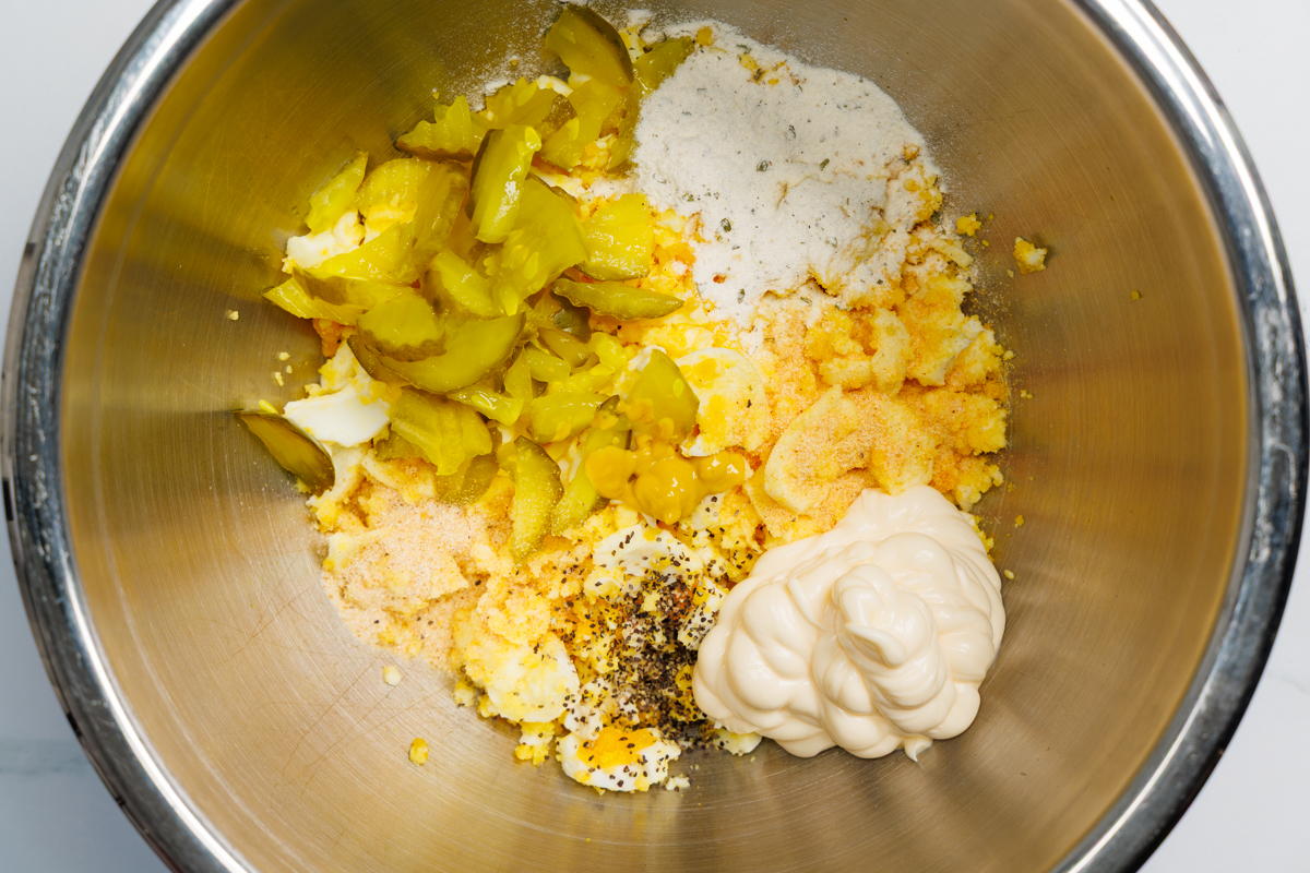 Chopped hard boiled eggs, chopped dill pickle chips, dill pickle juice, mayonnaise, yellow mustard, ranch seasoning mix, garlic powder, onion powder, and ground black pepper in a silver mixing bowl.