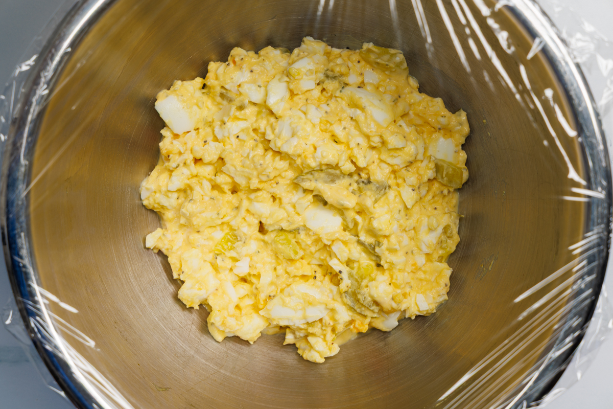 Dill pickle egg salad in a silver mixing bowl covered with clear plastic wrap.