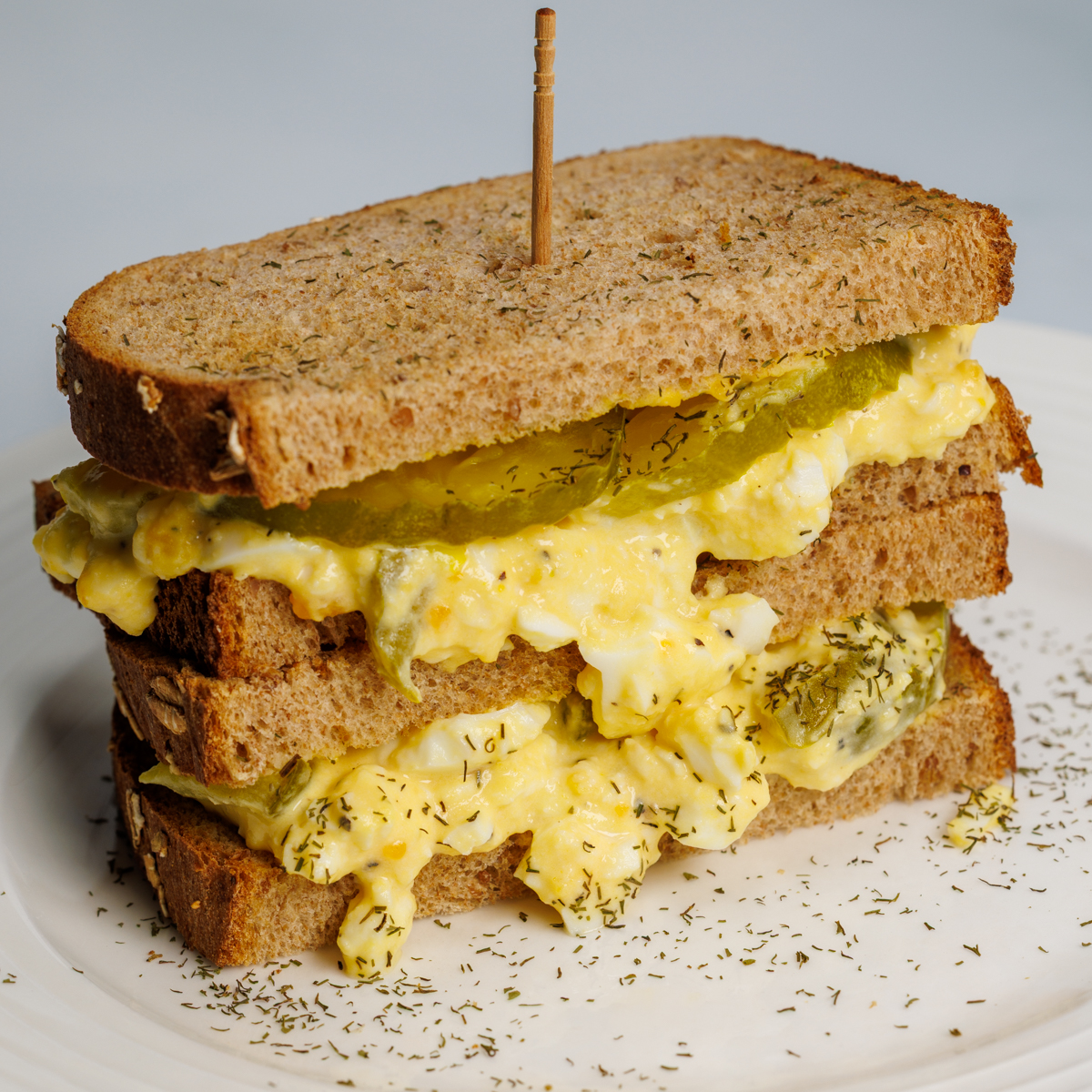 Two toasted whole wheat dill pickle egg salad sandwich halves stacked on top of each other with a wooden toothpick on a round white plate.