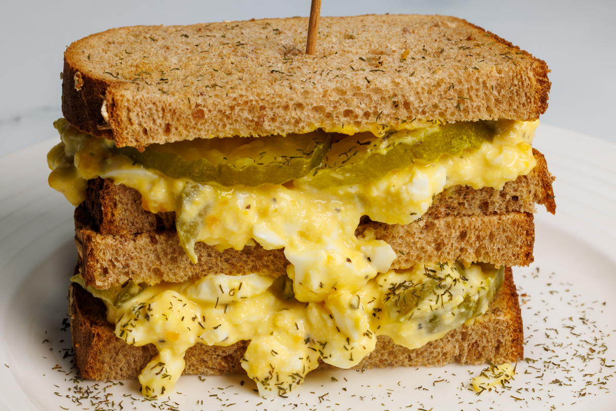 Dill pickle egg salad sandwich on toasted whole wheat bread cut in half and stacked on top of each other on a round white plate.